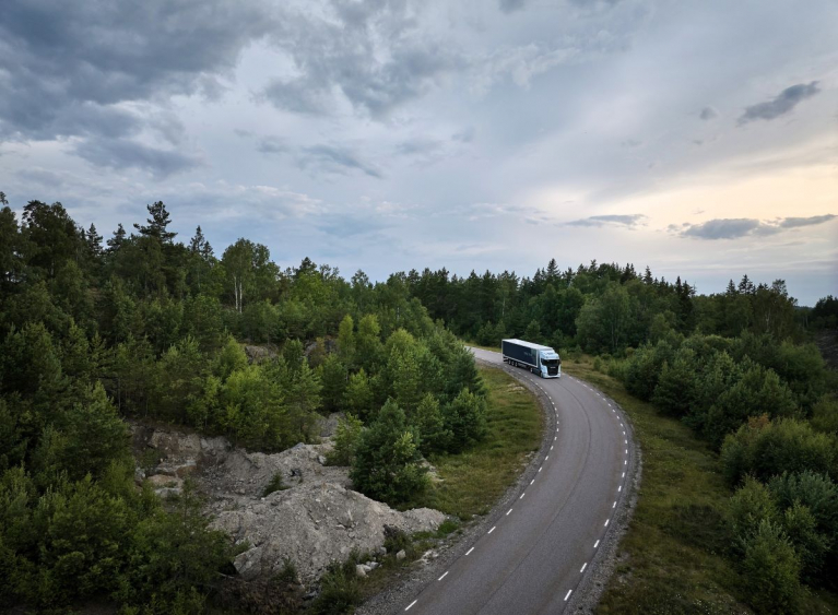 An electric Scania 40 R truck driving on a winding road through a lush green landscape under a cloudy sky.