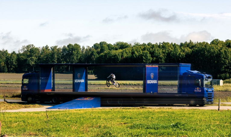 Two Scania autonomous trucks on a country road, going past each other in perfect sync to create a window of less than a second, allowing professional mountain biker and Red Bull Athlete Matt Jones to successfully jump between the two self-driving trucks.