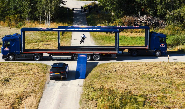 Two Scania autonomous trucks on a country road, going past each other in perfect sync to create a window of less than a second, allowing professional mountain biker and Red Bull Athlete Matt Jones to successfully jump between the two self-driving trucks.