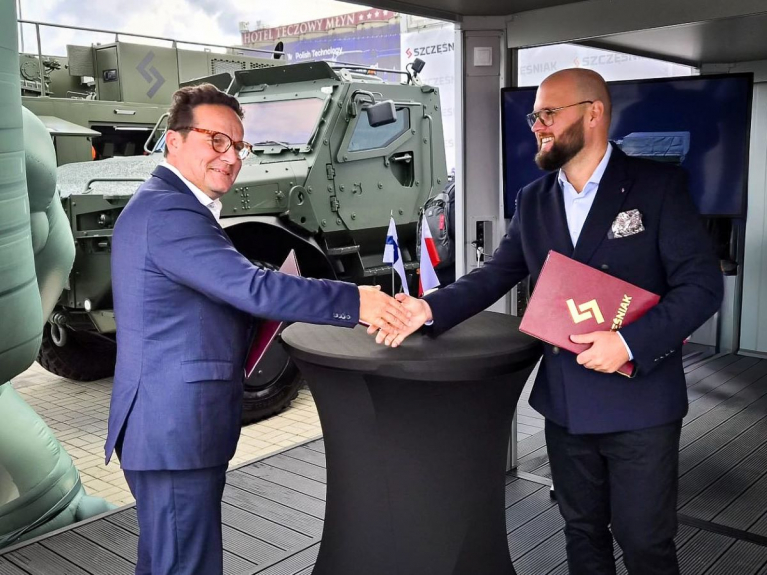 Sisu Auton ja Szczęśniakin edustajat kättelevät sopimuskansiot käsissään MSPO-messuilla. Taustalla on panssaroitu sotilasajoneuvo.