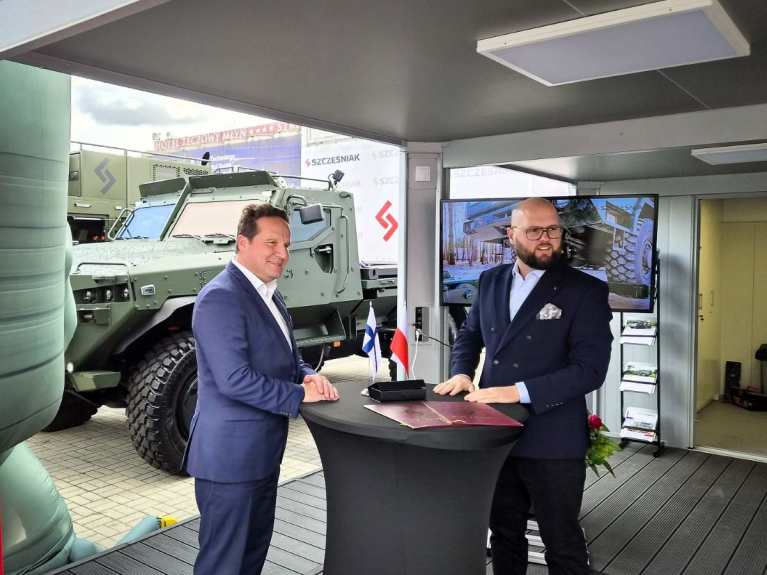 Sisu Auto and Szczęśniak representatives stand beside a table with agreement documents and Finnish and Polish flags at MSPO 2026, with an armoured military vehicle behind them.
