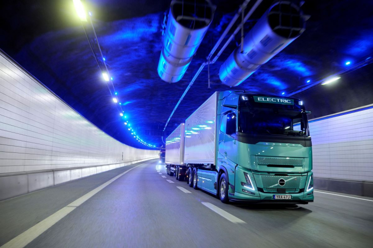 Green Volvo FH Electric truck with a box body and trailer driving through a tunnel illuminated by blue lights.
