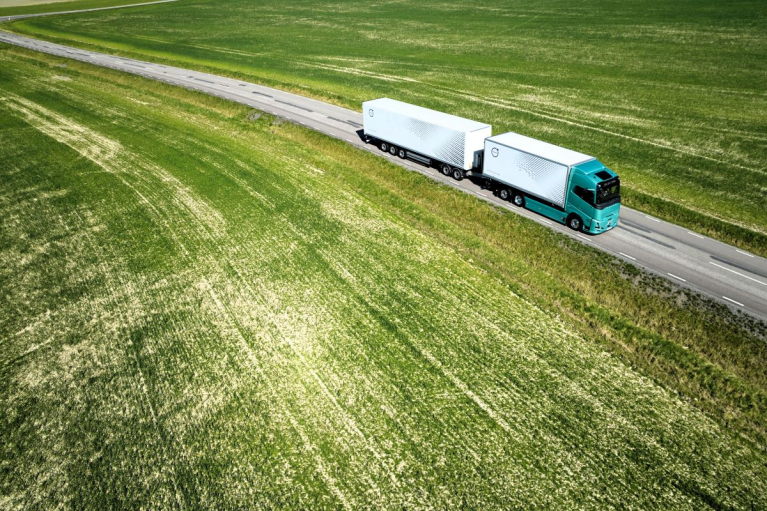 Ilmakuva turkoosista Volvo FH Electric -sähkökuorma-autosta ja valkoisesta umpikoriperävaunusta ajamassa peltojen halki kulkevalla maantiellä.