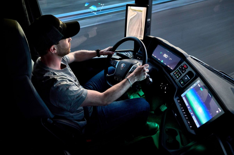 Driver at the wheel of a Scania truck, with a digital instrument cluster, central touchscreen and digital rear-view mirror display visible.