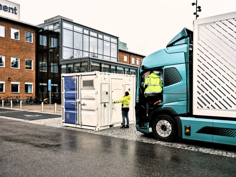 Volvo electric truck parked beside a containerised Volvo Penta battery energy storage unit, with two workers in high-visibility jackets.