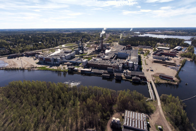 Ilmakuva Stora Enson Anjalankosken tehdasalueesta vesistön rannalla. Kuvassa näkyy tuotantorakennuksia, savupiippuja ja tehdasalueelle johtava silta.