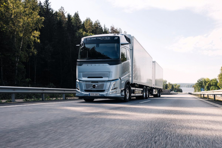 Gas-powered Volvo FH Aero truck with a box body and trailer driving along a forest-lined road.
