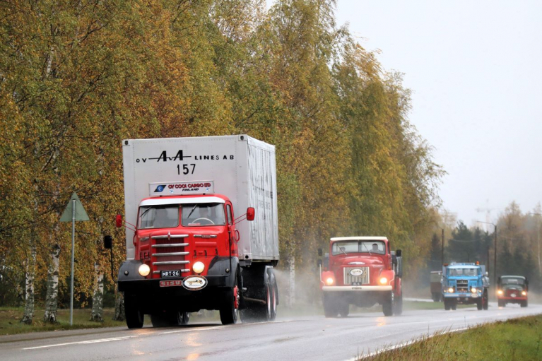 Punainen Sisu valkoisella umpikorilla johtaa vanhojen kuorma-autojen letkaa märällä, ruskapuiden reunustamalla tiellä.
