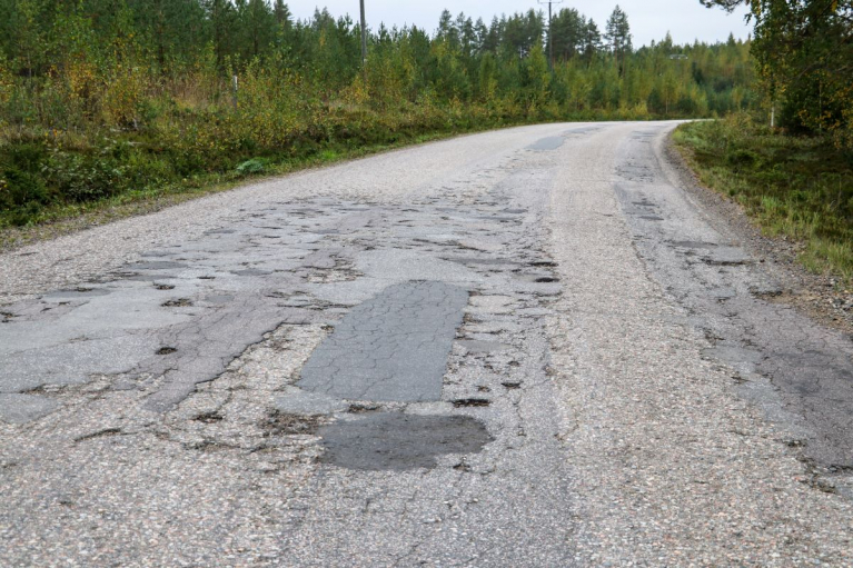 Metsän keskellä kaartuva päällystetty tie, jonka asfaltissa on runsaasti halkeamia, kuoppia ja paikkauksia.