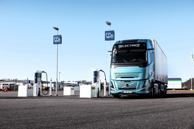 Turkoosi Volvo FH Electric -sähkökuorma-auto perävaunuineen raskaan liikenteen latausasemalla.