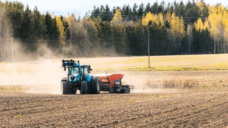 Sininen traktori vetää kylvökonetta pölyävällä pellolla. Taustalla näkyy syksyn väreissä oleva metsänreuna.