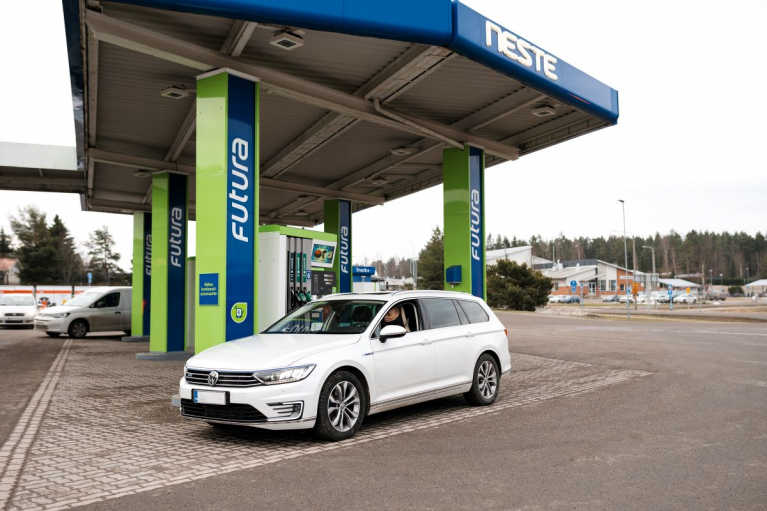 Valkoinen henkilöauto Neste-aseman polttoainemittareiden edustalla sinivihreän katoksen alla.