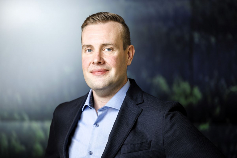Mikko Lintula tummassa pikkutakissa ja vaaleansinisessä paidassa metsäaiheisen taustan edessä.