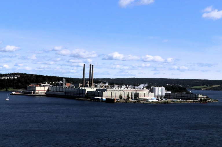 SCA’s Ortviken mill viewed across the water, with industrial buildings and tall chimneys against a blue sky.