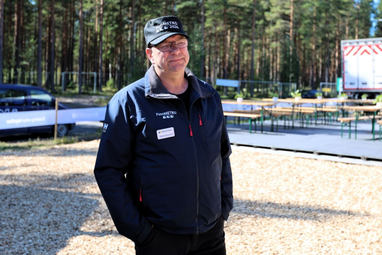 Koneyrittäjien puheenjohtaja Marko Vainionpää seisoo ulkona FinnMETKO-lippalakissa ja tummassa takissa. Taustalla on mäntymetsää ja pöytäryhmiä.