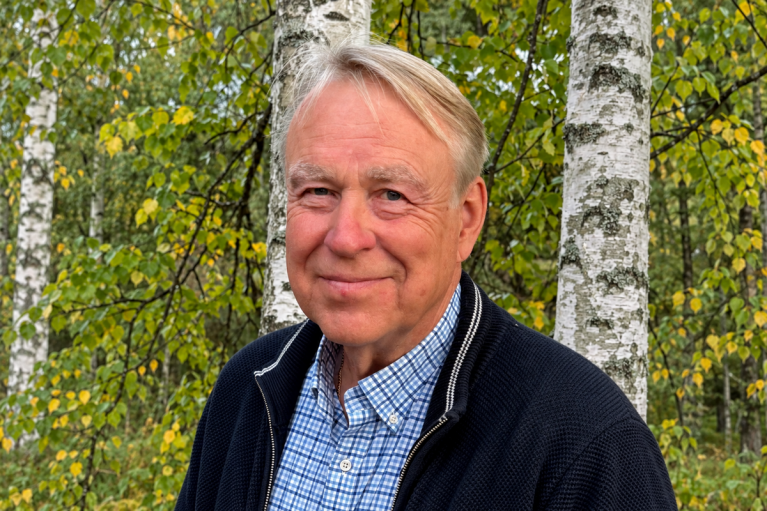 Keijo Ojala seisoo koivujen edessä yllään tumma neuletakki ja sinivalkoinen ruutupaita.