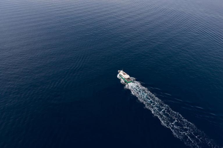 Aerial view of a boat crossing dark blue open water, leaving a long white wake.