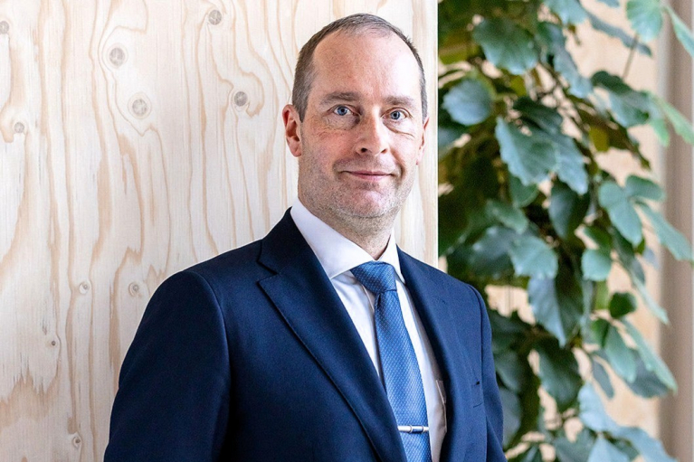 Bergslagets Skogar CEO Tuomas Hallenberg wearing a blue suit beside a wooden wall and a green indoor plant.