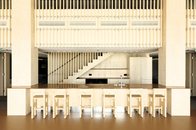 Light-coloured tables and chairs inside Keilaniemen Portti, with a staircase and timber balustrades in the background.